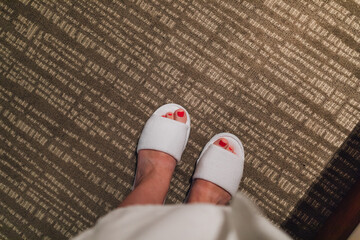 wearing a hotel terrycloth robe and looking down at feet with red pedicure in white spa slippers