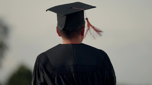 A man in a black graduation gown stands with his cap and gown on. He is looking off into the distance