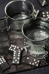 Zwei Glastassen in transparent grau – gefüllt mit Wasser – auf einem dunklen Untergrund mit Dominosteinen.