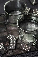 Zwei Glastassen in transparent grau – gefüllt mit Wasser – auf einem dunklen Untergrund mit Dominosteinen.