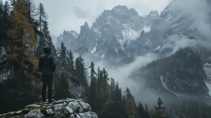 Chilly morning with solitary man standing on rocky edge amid dense forest facing rugged, misty mountain peaks in gray autumnal hues.