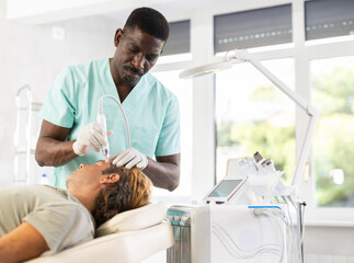 Male African cosmetologist performs skin cleansing procedure for European man client. Specialist...