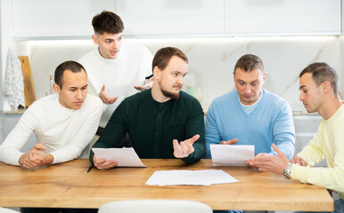 Upset worried man sitting at table in home kitchen with papers in hands, reading unexpected medical results with group of male friends solacing and comforting him ....