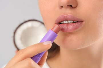 Young woman applying lip balm and coconut on light background, closeup