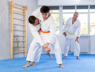 Two men practice grabbing and throwing sports mats during judo training under the guidance of an experienced elderly coach