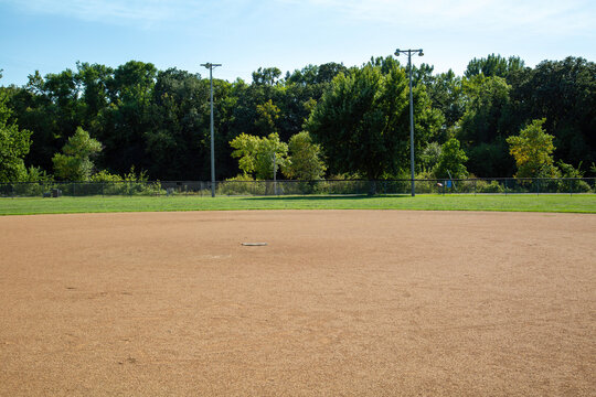 Empty Small Town Baseball Field Park under a Clear Blue Sky. Features Dirt Field, Green Grass, Chain Link Fence, Pitcher's Plate, and Distant Stadium Lights - Youth Community Sports and Recreation