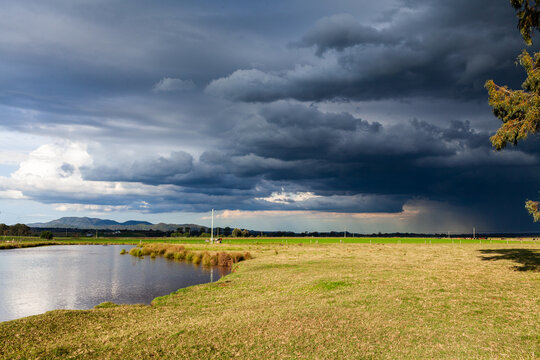 dark stormy rain clouds incoming over sunlit rural farm paddock and dam
