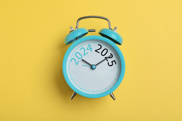 Alarm clock with hands pointing at numbers 2024 and 2025 on yellow background, top view. Beginning...