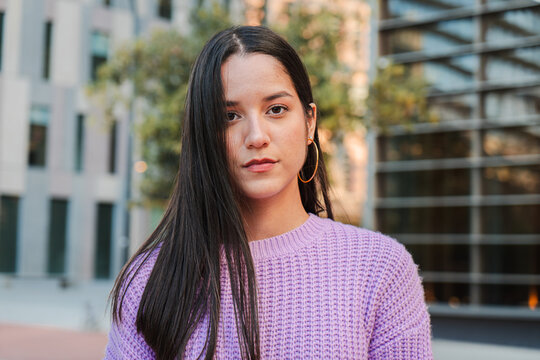 A Portrait of a Determined Young Latina female Student Standing Alone Outside, Gazing Forward with a Reflective Expression, Radiating Confidence and Self-Assurance. Woman looking serious at camera
