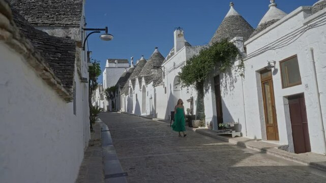 A young hispanic woman in a green dress walks down a cobblestone street in the charming old town of alberobello, italy, surrounded by iconic trulli houses under a clear blue sky.