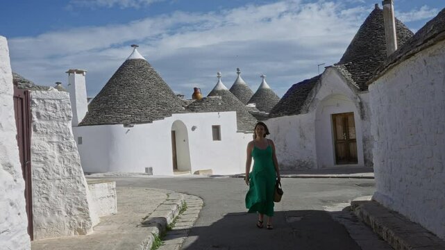 A young hispanic woman in a green dress strolls through the charming streets lined with iconic trulli houses in the historic town of alberobello, puglia, italy, on a sunny day.