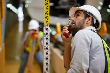 Worker or engineer using theodolite surveying optical and coworker holding large ruler for...