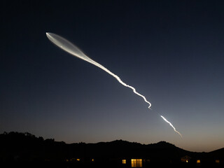 A bright white trail from a rocket launch stretches across the night sky as it ascends into space. The trail is visible against a dark blue sky, with a line of silhouetted hills.