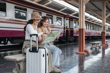 Two young Asian women sitting on a bench at a train station, holding a map and pointing, with a...