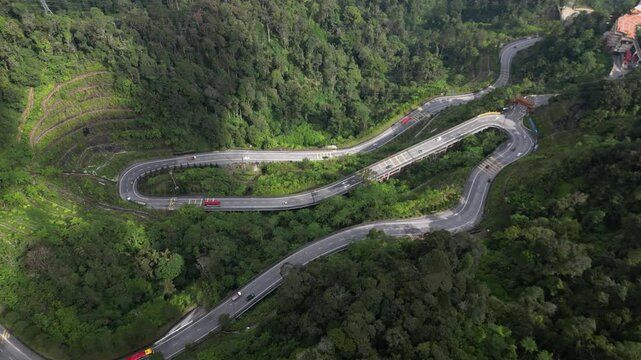 Aerial view of a serpentine road meandering through a dense green forest with vehicles driving along. rising shot