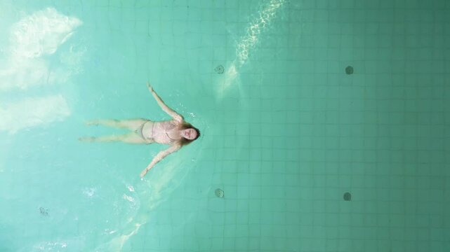 Female swims on back kicks with extra wide leg movements in pool, overhead drone