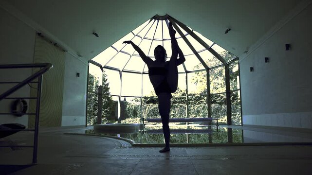 Silhouette of flexible female gymnast next to indoor pool doing leg hold