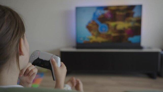 A 7-year-old female child intensely playing computer games on a PlayStation, captured from the back. This stock video highlights the focus of a young gamer, illustrating modern childhood.