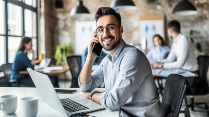 Smiling businessman talking on phone while working on laptop in modern office with colleagues in background.
