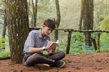 Fototapeta premium Portrait of confident Asian man writing on notebook while thinking and looking away in the nature forest. Peaceful and relaxing healthcare wellness