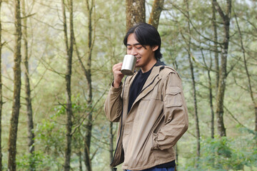 Portrait of excited Asian hiker man drinking water from stainless steel cup while trekking in nature forest