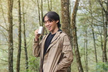 Portrait of excited Asian hiker man drinking water from stainless steel cup while trekking in nature forest