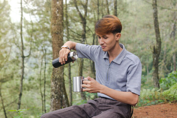 Portrait of excited Asian man pouring water from the vacuum flask to the stainless steel mug. Drinking mineral water while trekking in nature forest