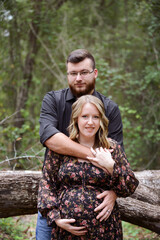 A pregnant couple embracing in a forest, with the man's arms aro