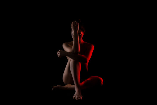 The image shows a woman sitting in a dark studio with red light illuminating her body. She is facing the camera with her hands clasped together near her chest, and her head is slightly tilted down