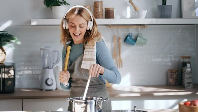 Video of funny happy young woman listening to music with headphones while cooking raw spaghetti in the kitchen at home