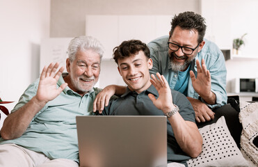 Video call concept. Happy multigenerational family sitting on sofa using laptop technology for webcam connection - grandfather with mature son and young grandson enjoying carefree moments together - Powered by Adobe