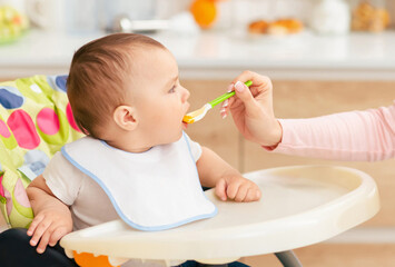 A mother is feeding her baby boy who is seated in a high chair in the kitchen. The atmosphere is...