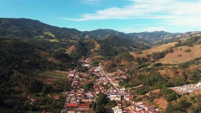 Aerial footage of Delfim Moreira, a charming city nestled in the mountainous region of Minas Gerais, Brazil. The city's red-roofed buildings surrounded by lush green hills under a bright blue sky