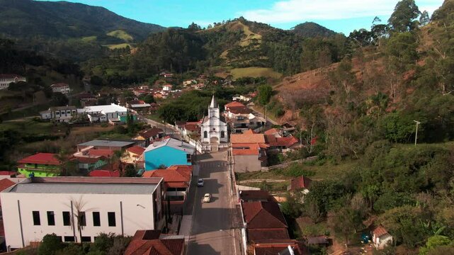 Aerial footage showing the central area of Delfim Moreira in Minas Gerais, Brazil. The city is surrounded by green hills and features a prominent church and colorful buildings under a bright blue sky