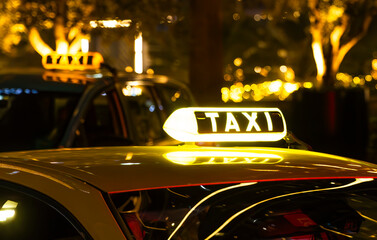 A yellow taxi cab with a lit-up TAXI sign on the roof. The cab is parked in a city at night, with blurry lights in the background.