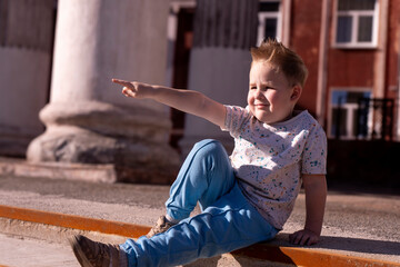 A cute little boy sits on the steps among the columns of an old building and points his finger...