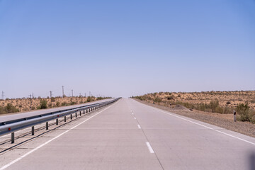 A long, empty road with a blue sky above. The road is lined with metal guard rails