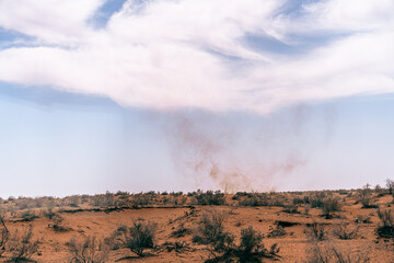 Fototapeta premium A cloud of dust rises from the desert floor. The sky is clear and blue, with a few clouds scattered throughout. The scene is desolate and barren, with only a few sparse trees in the distance