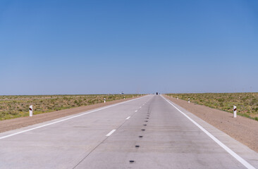 A long, empty road with no cars or people. The sky is clear and blue. The road is made of concrete and has white lines painted on it