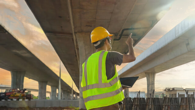 Asian construction supervisor in yellow vest and hard hat uses laptop to inspect pointing at the bridge This scene takes place on a highway construction site.