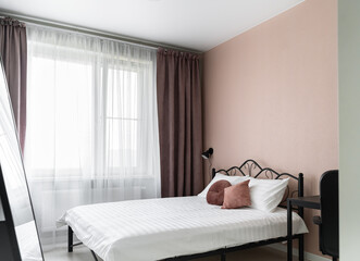 design of bedroom with beige walls, a big bed with a metal headboard, a white bedspread and pillows. There are flowers in a vase on the table in the bedroom, a chest of drawers and a mirror