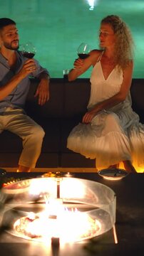 Romantic couple by poolside fire pit, toasting with wine, enjoying cozy atmosphere