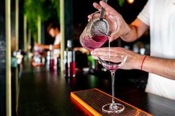 A skilled bartender pours a vibrant pink cocktail into an elegant glass at a dimly lit bar, showcasing the art of mixology