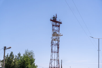 A tall tower with a satellite dish on top. The sky is clear and blue. The tower is surrounded by trees