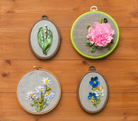 Four types of spring garden flowers are embroidered with satin ribbons in hoops on wood background. Daisies, pansies, lilies of valley and peony. Embroidery as hobby and leisure. Gifts for Mothers Day