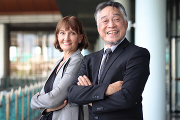 Happy smiling senior Asian businessman in suit with Caucasian businesswoman standing with arms crossed at office. Elderly successful male and female boss at workplace. Portrait  mature businesspeople.