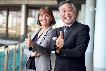 Happy smiling senior Asian businessman in suit with Caucasian businesswoman giving thumbs up to camera at office. Elderly successful male and female boss at workplace. Portrait mature businesspeople.