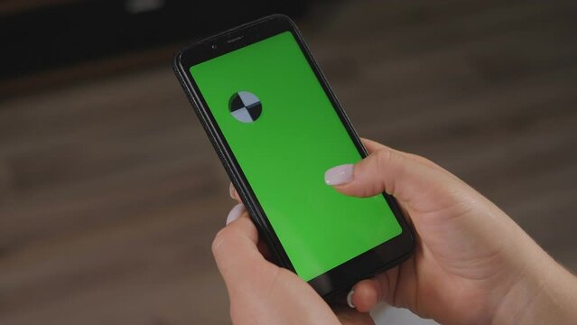Close-up of woman's hand browsing smartphone with green screen at home. Woman at home relaxing reading on the smartphone with pre-keyed green screen.