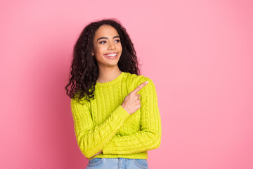 Fototapeta premium Portrait of beautiful curly haired brunette beginner saleswoman in green knitted pullover pointing finger promo isolated on pink color background