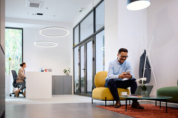 Happy businessman texting on smart phone while working on laptop in office.